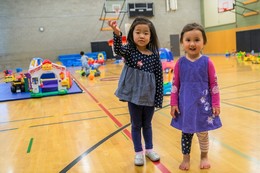 Two toddler girls are standing in a gymnasium with toys