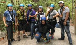 A group of youth stand outside in a wooded area with climbing gear on.