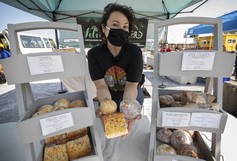 A vendor wearing a face mask holds pastries for the camera while standing at a farmer's market booth.