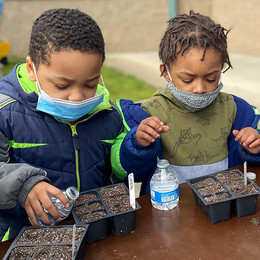Two children in coats stand at a table and water newly planted seeds in six-pack seed pots.