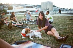 A woman with a dog sitting on grass in a park with other people and bikes around her