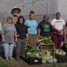 A group of people standing in front of a large greenhouse with crates of vegetables displayed in front of them. 