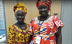Two women in African print head dresses and dresses stand together smiling