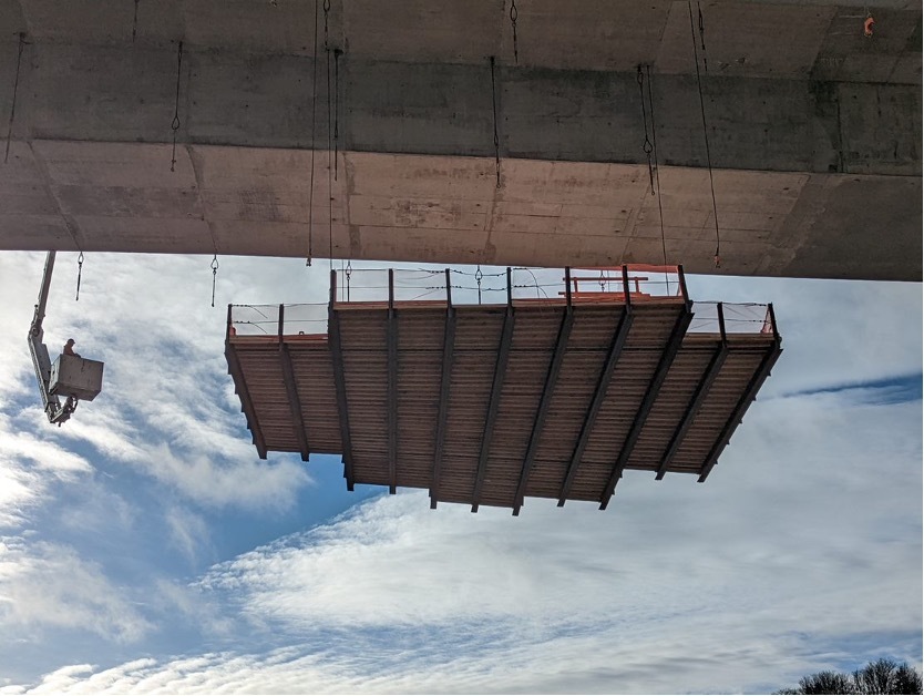 Using winches positioned on the bridge deck, the platform is lifted from the ground to the underside of the bridge. 