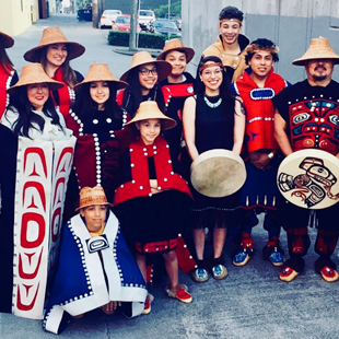Group of people in traditional Haida dress smiling and holding drums on a Seattle street.