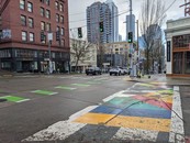 Intersection with traffic lights, painted bike lane and colorful crosswalk with buildings in the background and cars. 