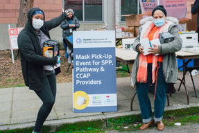 Two child care providers smile for a photo at DEEL's mask giveaway event with boxes of PPE supplies in their hands.