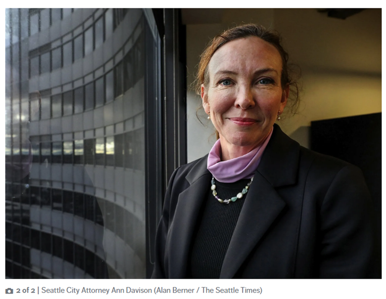 City Attorney Davison looks directly at the camera and smiles with building windows in the background