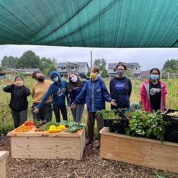 A group of teenagers stand in front of raised garden beds displaying recently harvested produce