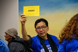 An Asian man wearing a blue shirt holding up a yellow sign that reads: "Assist."