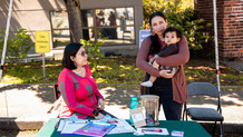 Two Native women stand at a table with informational forms, one of them holding a young child