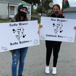 Two youth stand holding signs that "Safe River, Safe Salmon" in English and Spanish