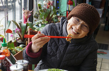 An Asian woman smiling and holding up a piece of tofu in a pair of chopsticks 