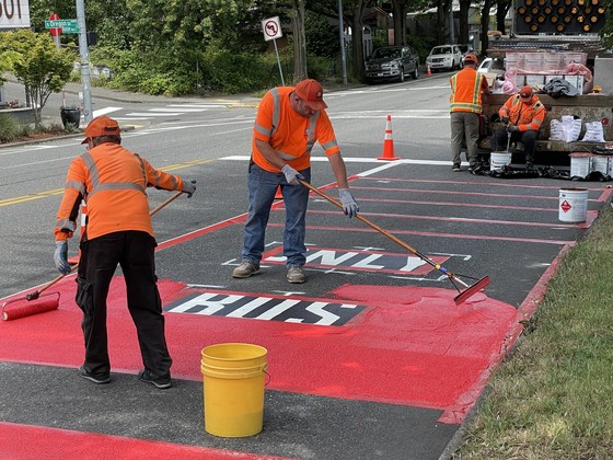 Construction workers wearing brightly colored vests apply red paint to the street in a lane labeled "bus-only" 