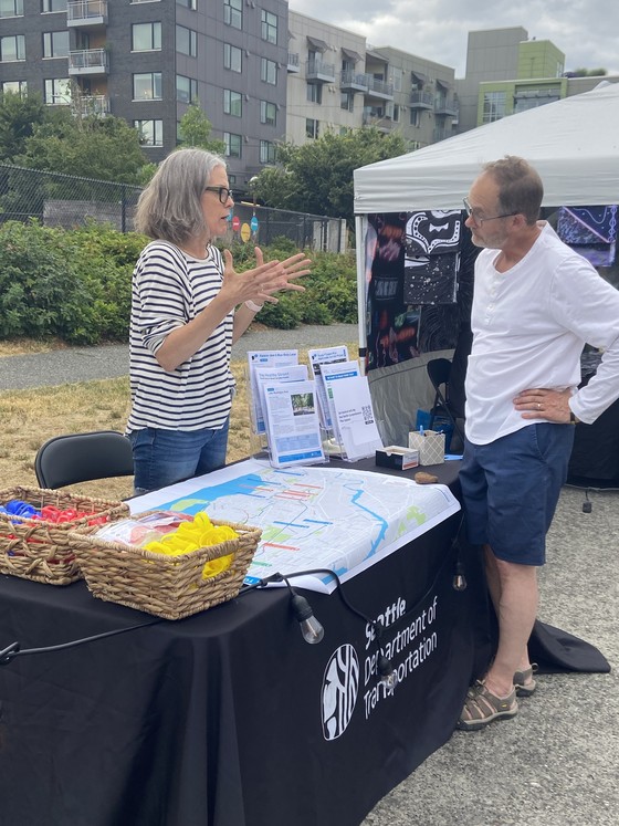 Woman speaks to a man at a table that contains information boards, papers, and other project information