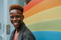 Photo of smiling Black youth standing in front of a wall painted with rainbow stripes [Photo by Anete Lusina on Pexels]