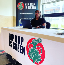 A Black woman sitting behind a desk with signage that reads "Hip Hop is Green"