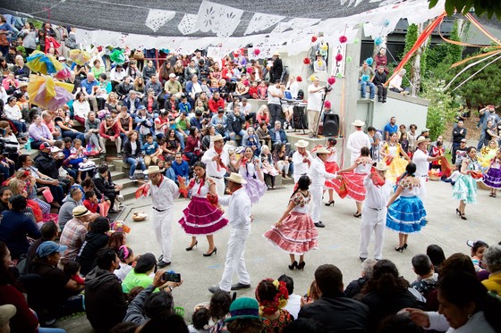 A group of Latinx dancers performing in traditional dance attire while a crowd watches in a park amphitheater stage.