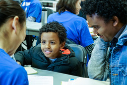 A melanated, smiling small child with dark, curly hair is sitting next to his father and talking with a citizenship clinic volunteer.