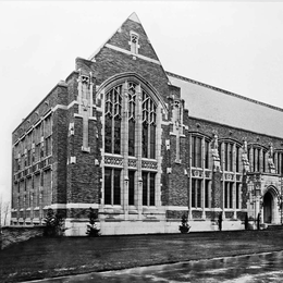 A black and white photo of a brick building with gable roof and large windows
