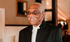 An older Black man smiling, and wearing a black suit jacket, white shirt, and glasses