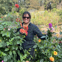 A young, Indian woman wearing sunglasses, smiling, standing in a bed of dahlias 