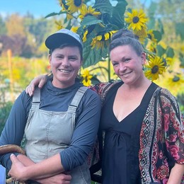 Two woman standing in a community garden with a large bunch of sunflowers behind them.