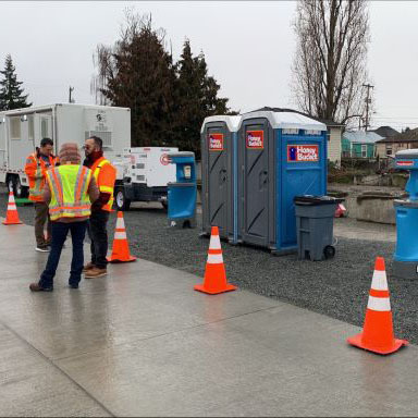 A group of emergency response workings talking in front of a set of porto potties and construction cones
