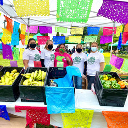 A group of people standing behind a table with crates of produce and colorful flags hung all around the background.