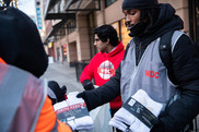 A Black man dressed in warm clothes and a gray vest with red letters "WDC" hands out packs of socks with a brick building in the background.
