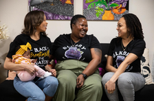 Three Black women, one holding a baby, sit on a couch looking at each other with large smiles. 