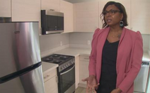 A Black woman stands in a kitchen wearing a pink/red blazer and speaking to someone off camera.