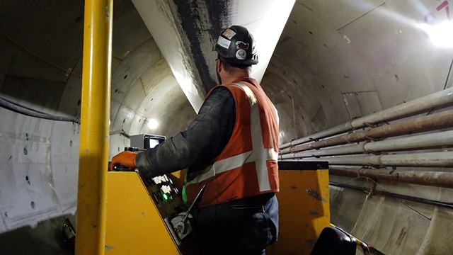 Person in orange vest and yellow hard hat stands on yellow construction equipment in tunnel, facing away from camera