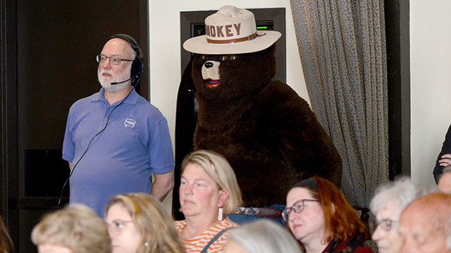 Man in blue polo, headset, and white beard stands next to Smokey Bear behind seated crowd