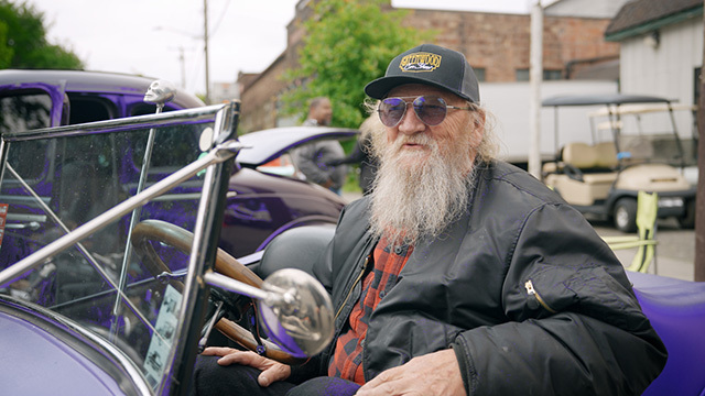 Bearded man in black baseball cap and sunglasses sits in purple convertable