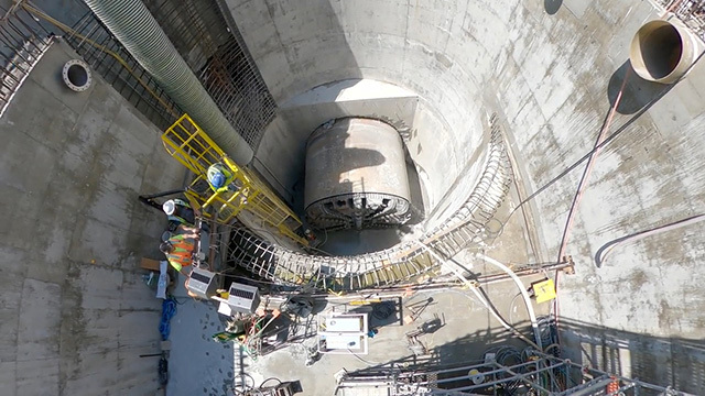Three construction workers stand on equipment in cylindrical cement pit