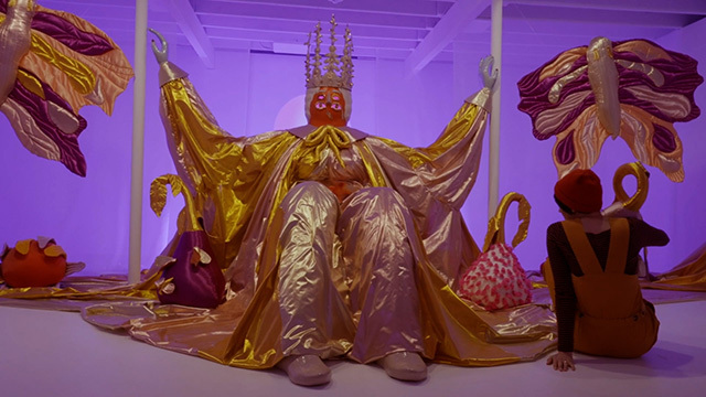 Person in red beanie sits with back to camera in front of art humanoid art piece wearing crown and gold and silver robe