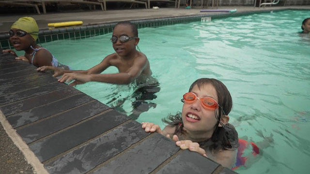 Three kids in goggles hold onto the edge of a pool