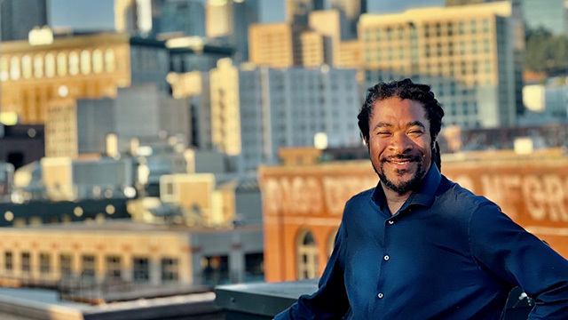 Black man in blue shirt smiles in front of blurred city background