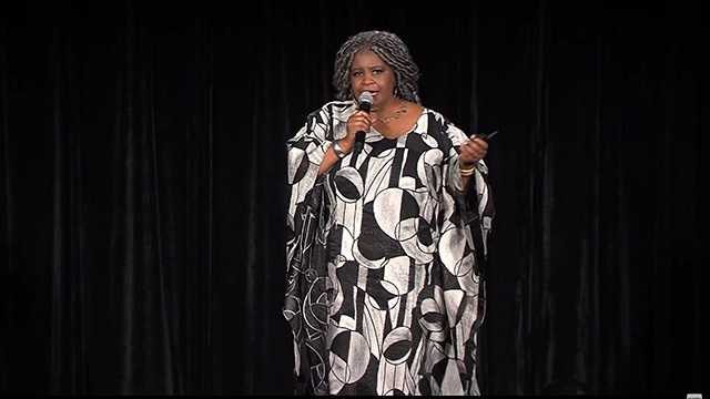 Black woman in long gray and white dress holds microphone onstage