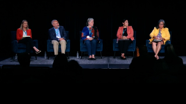 Five people in matching chairs sit onstage in front of black background looking at woman on far right in yellow blazer