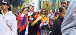 An Indigenous woman wearing traditional attire and playing a drum walking down the street with a group of people