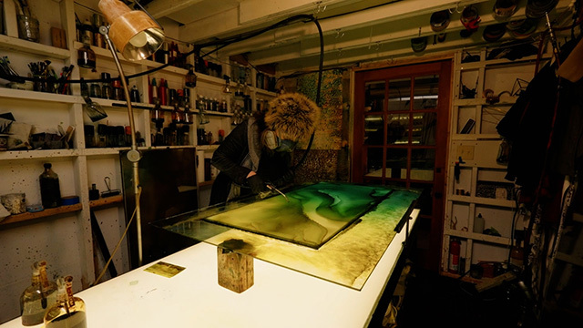 Person in large fuzzy hat and black clothing covering almost all skin in dim art studio leans over light table with green painting