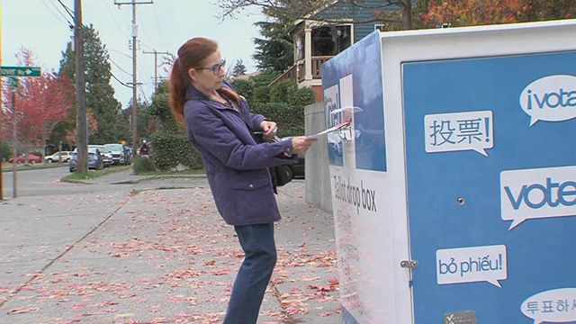 Person with long red ponytail, purple jacket, blue jeans puts ballot into ballot box.