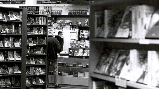 Black and white image of video store shelves, one person back to camera across from man behind camera
