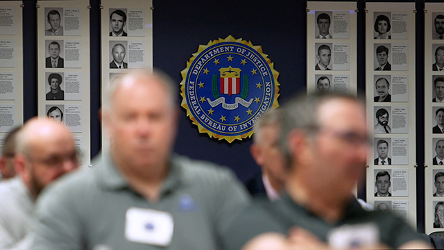 Blurred people in foreground, blue wall with black and white photos and FBI seal behind