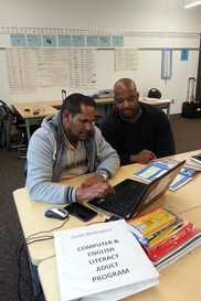 A teacher watches a man perform a task on a laptop in a classroom