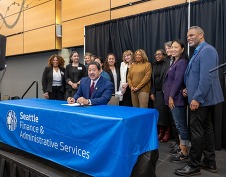 Mayor Harrell seated at a table with a banner reading Seattle Finance & Administrative Services with staff and business leaders standing behind him