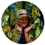 Someone peeks out from lush hanging vines smiling and framing their face with their hands.