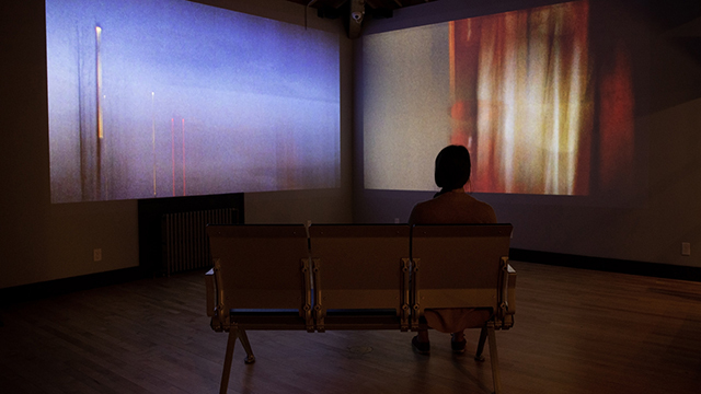 View of person sitting in row of three theater seats from behind facing light art on two walls 